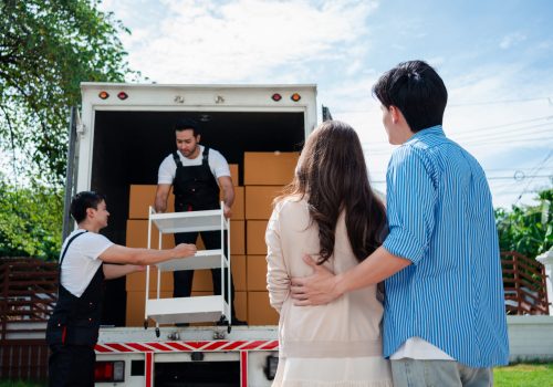 Asian Couple check while unloading boxes and furniture from a pickup truck to a new house with service cargo two men movers worker in uniform lifting boxes. concept of Home moving and delivery.